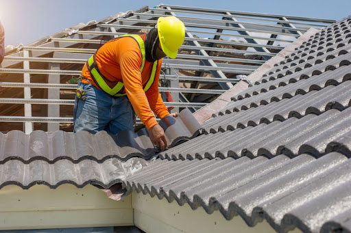 A roofer installs part of a residential roof. He is wearing a hardhat and safety vest. You can not see his vest as he is looking down. He is at an angle on the roof structure.